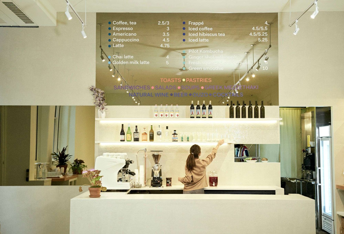 A woman reaches up for a glass on a shelf behind a bright white counter. On the counter is a potted plant, a large espresso maker, and a coffee maker. A large wall menu sits above the shelf.