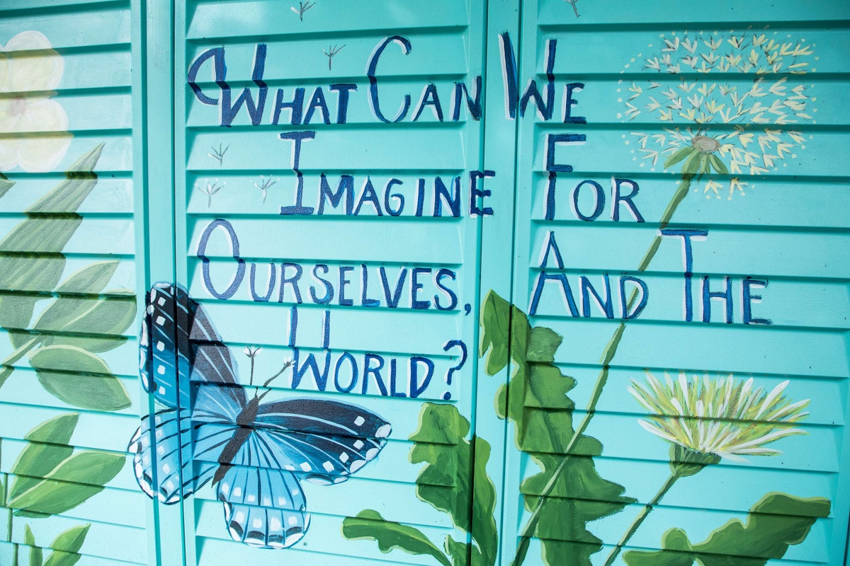 Blue shutter doors with butterfly and dandelion mural reading "What can We Imagine For Ourselves, And World?"