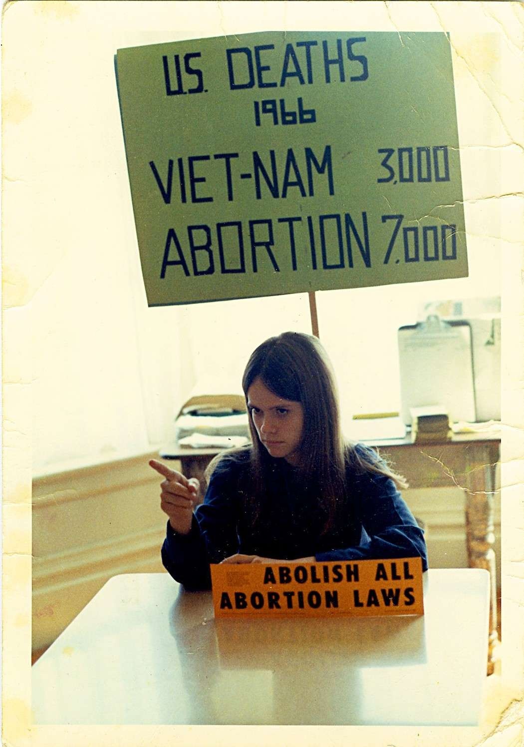 A young abortion rights activist sitting at a table with a sign behind her that reads: "U.S DEATHS, 1996, VIETNAM: 3,000, ABORTION, 7,000"