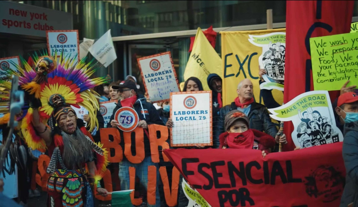 Protesters hold a variety of red, yellow, green, and orange signs written on various materials such as cloth, nets, and paper. One person stands out amongst the crowd wearing a colorful headdress.