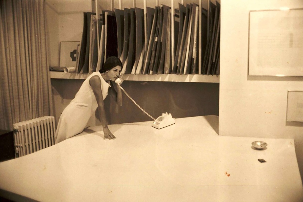 Sepia toned image of gallerist Paula Cooper leaning on a white desk on a telephone at Park Place Gallery