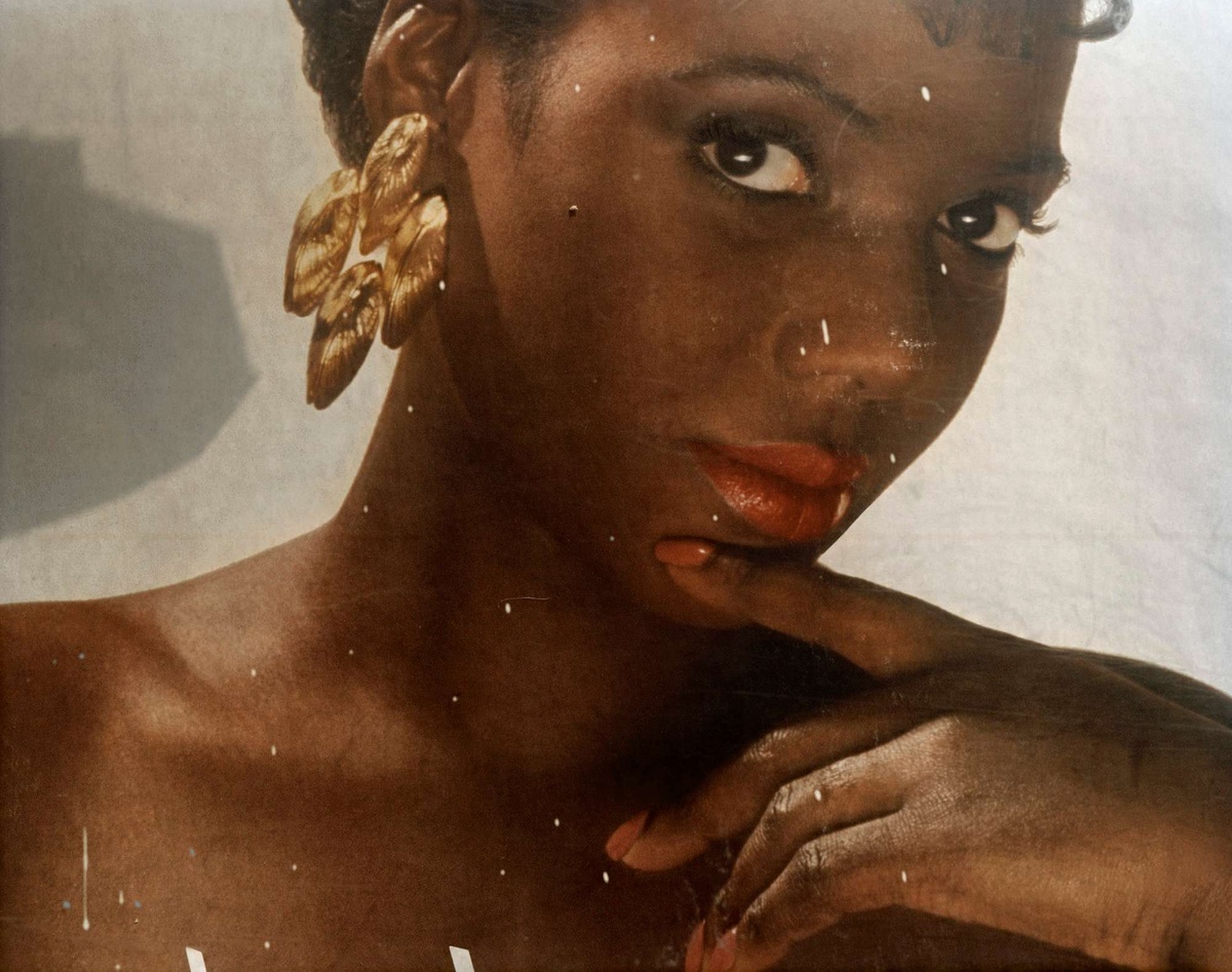 A slightly damaged photograph of a Black woman facing the camera at a 3/4th angle. She is wearing glam makeup; a smokey eye, skinny brows, and red lips. She has a large, shining gold earring. Her hand rests just below her lip with red nail polish that matches her lip color.