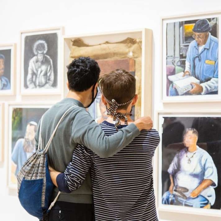 Two people embrace while looking at a wall of framed artworks