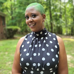 C.M. Lockhart, a Black woman with green hair smiling with a forest backdrop behind her