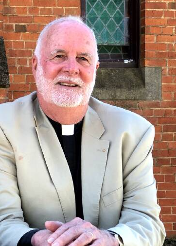 The Venerable Dr John C. Davis, wearing a black shirt and white jacket. He is smiling and looking towards the camera. In the background is a redbrick wall and the lower portion of a lead light window