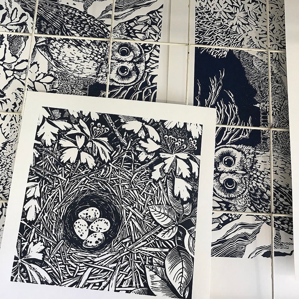 black and white lino print of a bird's nest nestled in leaves on a drying rack. Prints of owls can been seen in the rack below.