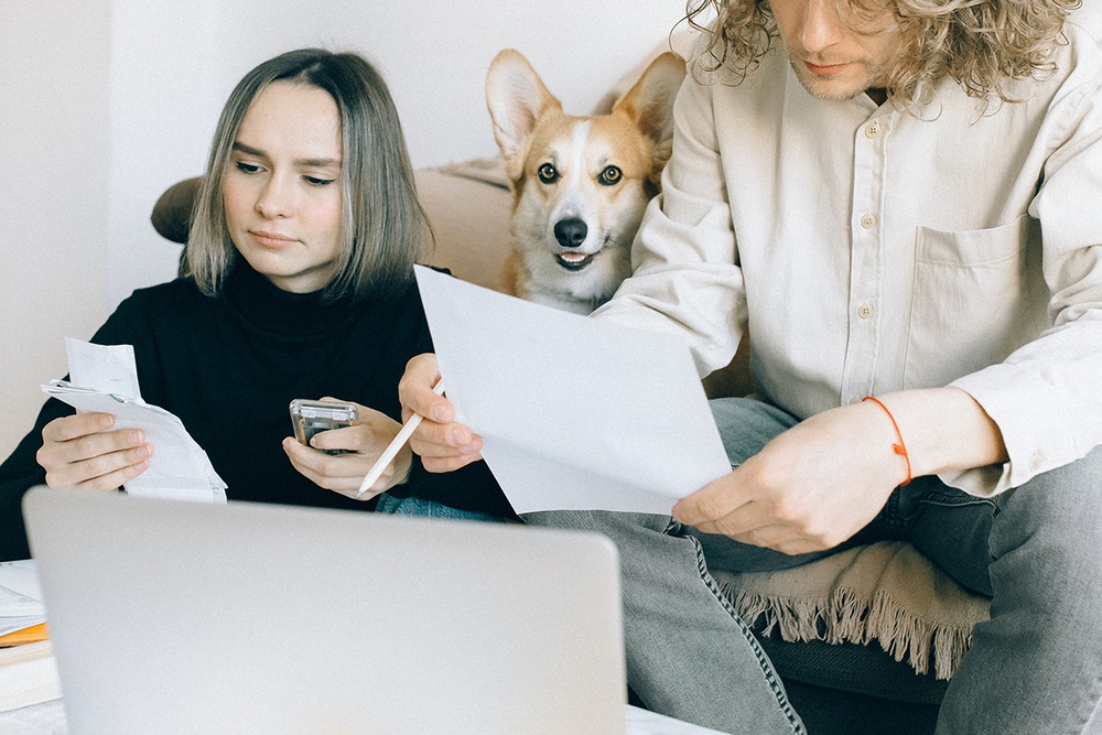 A young couple review papers and receipts in front of their open laptop while comparing pet health insurance plans while a corgi sits between them unbothered