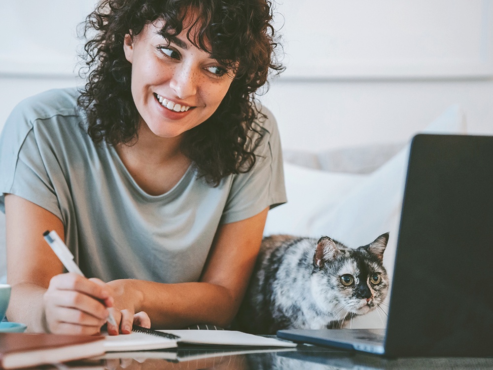 A brunette woman takes notes at her virtual veterinarian appointments while her gray and black cat looks at the laptop screen