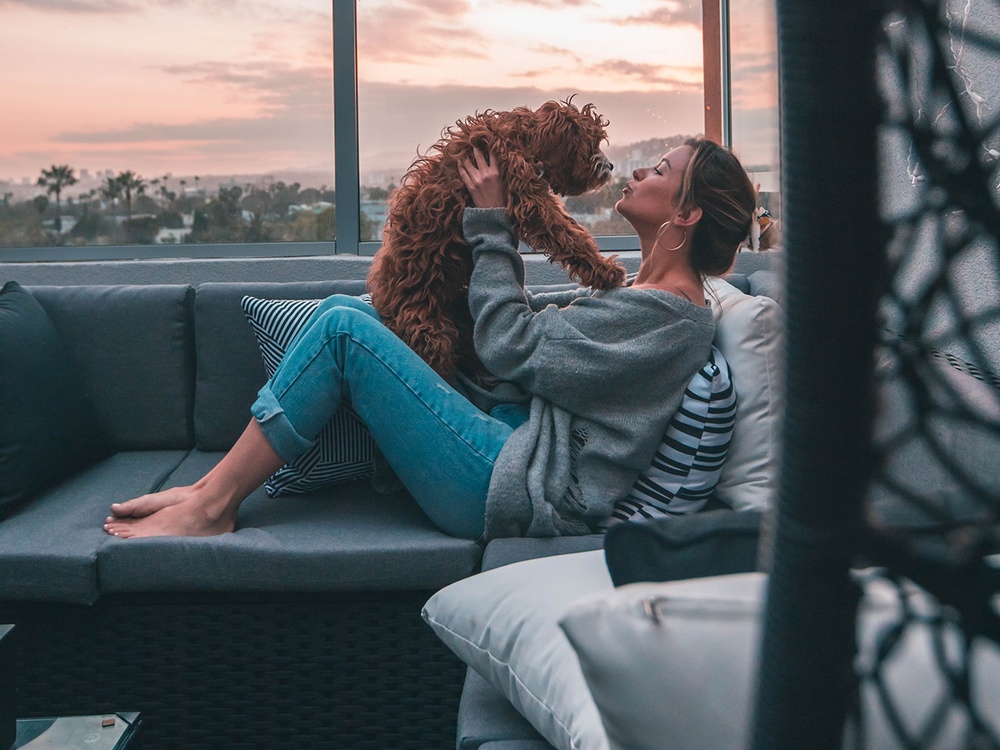 Sitting outside on a rooftop patio, a woman in casual clothing holds a goldendoodle up while making a kissing face