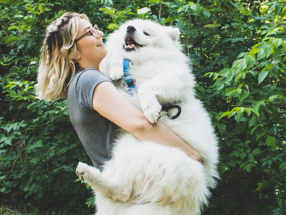A blonde woman holds a giant samoyed dog in her arms in front of a solid wall of green vines.