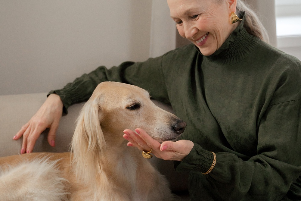 An older woman in an emerald green dress and prominent gold jewelry gently pets her older large dogs face while smiling
