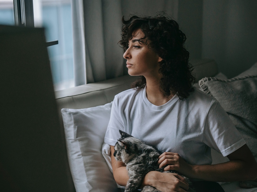 A brunette woman looks out the window worriedly while holding her black and white cat. She wonders when should I bring my pet to the emergency vet? 