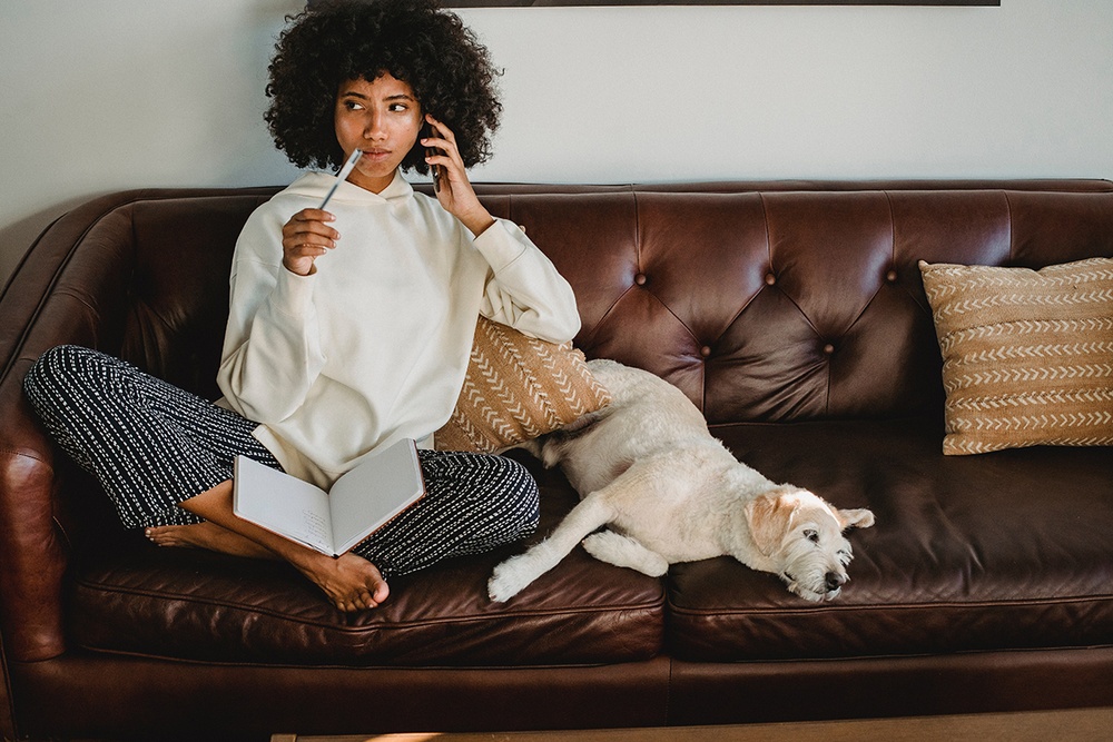 A woman sits, unsure on her leather sofa while her white medium size dog lays next to her.