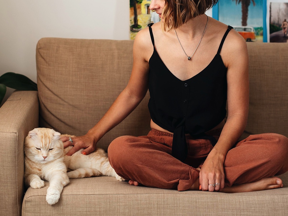 A blonde woman sits crossed legged on a tan sofa while reaching out to pet the cream cat laying next to her. 