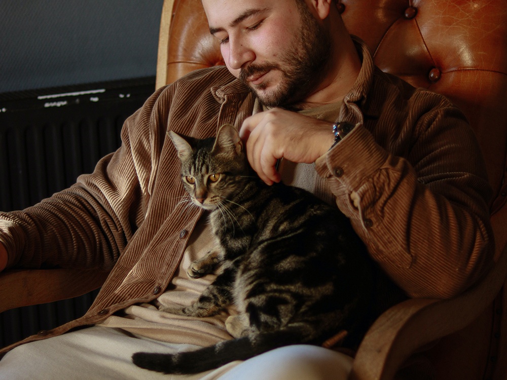 A tabby cat lays on a man in a corduroy jacket, He gently pets her shoulders.
