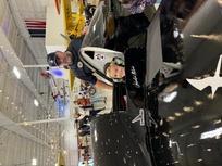 A young child with a big smile sits in the cockpit of a sleek black aircraft, wearing headphones and looking excited. A man standing behind the child, wearing a navy shirt and cap, looks on proudly. The event is indoors with other people and aircraft in the background, creating a vibrant and engaging atmosphere.