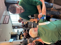 A man in a green jacket is practicing first aid on another man's arm, applying a blue tourniquet. They are seated at a table in a large indoor space with other people nearby. The setting appears to be a group training or workshop, with participants actively engaged in learning. A sign for the Delta Sky360 Club is visible in the background.