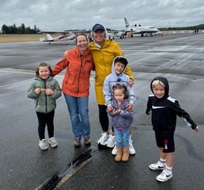 Two smiling women, one in an orange jacket and the other in a yellow raincoat, stand together with a group of children on an airport tarmac. The children, dressed in rain jackets and hoodies, are happily posing for the photo. Several small airplanes are visible in the background under a cloudy sky, setting the scene for a fun, family-friendly day at the airfield.