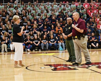 3 people talk on gym floor with crowd in bleachers