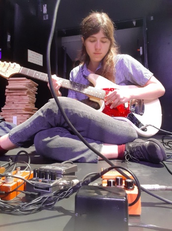 Maya Perry playing an electric guitar while seated on the floor with pedals and wires in the foreground.