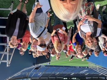 A large group of smiling individuals gathers on the field at a stadium. Many are holding papers, and they appear to be enjoying a tour or group activity. The scene takes place in bright daylight, with the green turf and the empty stands of the stadium visible in the background. The group is standing close together, creating a sense of excitement and camaraderie.