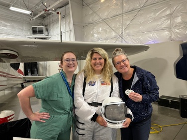 Three women are smiling and standing together in front of a small aircraft in a hangar. The woman in the center is dressed in a white astronaut suit, holding a space helmet. The other two women, one in a green jumpsuit and the other in a blue jacket, are also smiling and holding a commemorative patch. The environment is lively, with aviation-related activities happening around them.