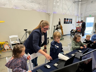 A woman wearing glasses and a navy jacket is smiling while assisting a group of young children with computers at a Minecraft Coding e-sports table. The children are focused on the laptops, engaging in coding activities. The scene takes place in an indoor hangar, with various aviation-themed equipment visible in the background.