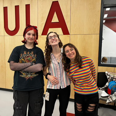 Three smiling individuals pose together in front of a wooden wall with large red letters spelling "ULA." The person on the left has bright red hair, a black t-shirt, and fishnet sleeves. The person in the middle wears glasses and a striped shirt, while the person on the right wears glasses and a colorful, rainbow-striped sweater.