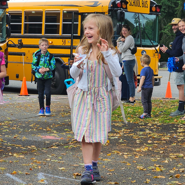 A young girl, smiling with excitement, walks toward the camera in a striped dress while arriving at school. She is surrounded by other children and adults, with yellow school buses visible in the background. The scene captures the joy of a school day filled with energy and smiles.