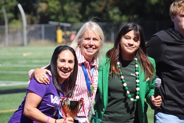 Three women stand together on a large football field. The left woman is brunette, wearing a purple shirt, smiling and holding a small trophy, the middle woman is wearing a striped blouse, a gold medal and has her arms around the other two, the third young woman is in all green smiling and holding a microphone