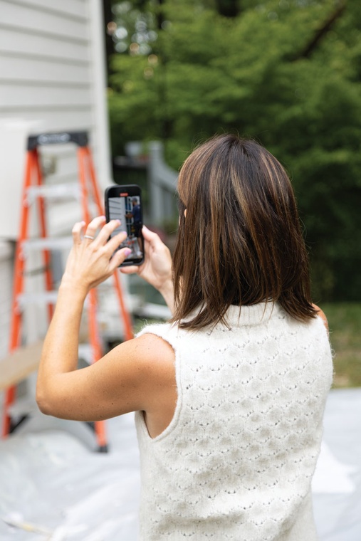 A brunette woman uses her phone to record video of window installers working on her home.