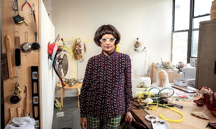 Rina Banerjee in the center of her studio with equipment hanging on the left and a table with materials on the right.