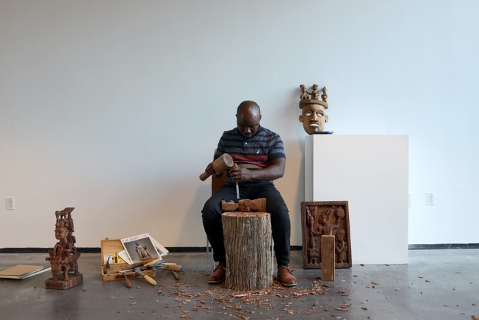Lukman Àlàdé Fákẹ́ye sits in the center surrounded by Yoruba sculptures, using a block and chisel to create a new work atop a tree stump.