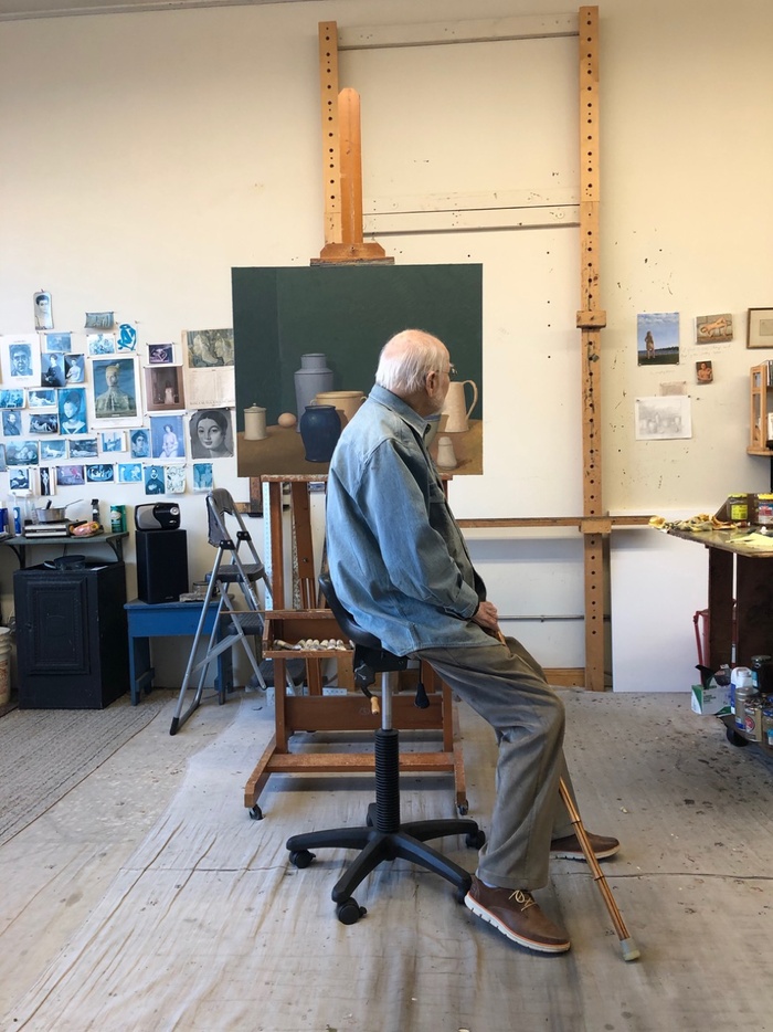 A still image of painter and professor William Bailey sitting in his studio in front of a work-in-progress on an easel.