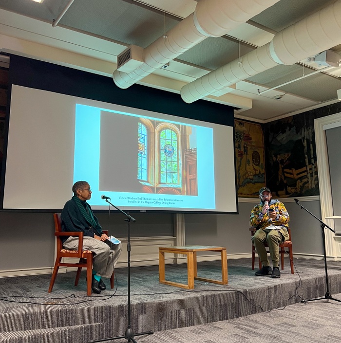 Barbara Earl Thomas sits opposite Christopher Paul Jordan on a small carpeted stage with a projection of a photo of Thomas’ stained glass commission behind them.