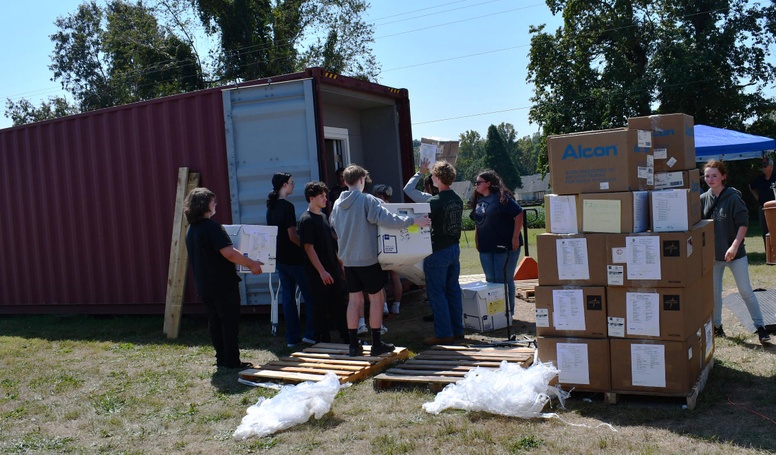 9 people carry boxes into shipping container with stack of boxes nearby