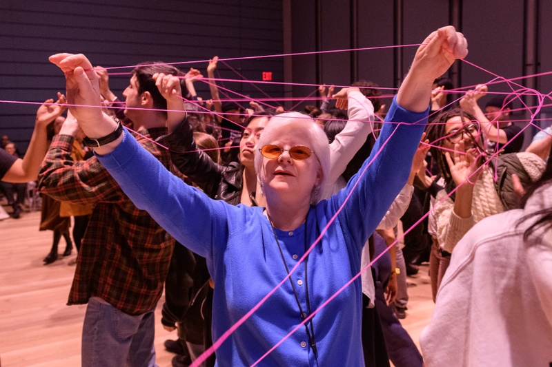 About 100 people stretching their arms while holding pink strings, creating a web of connections. Inside of a large theater.