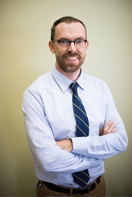 James McKowen, Ph.D. - a white person wearing a button up shirt and tie smiles at the camera while standing with his arms crossed across his chest.