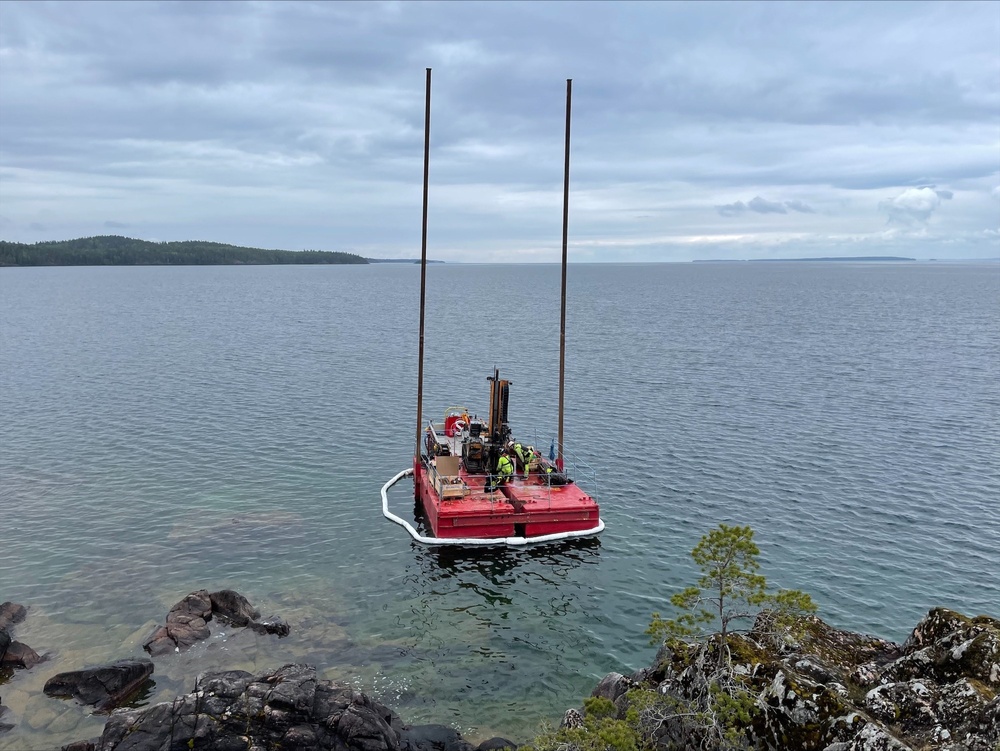 En röd pontonliknande flotte syns ute i vattnet nära land i norra Vättern. På flotten syns en borrigg samt personal (två personer) iförda gula skyddskläder.