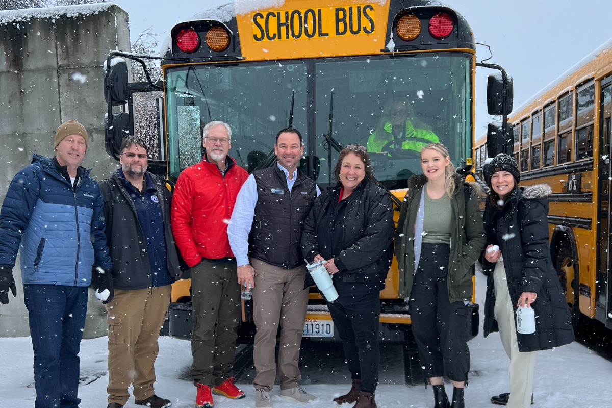 A group of people stand in front of a school bus in the snow, smiling for a photo.