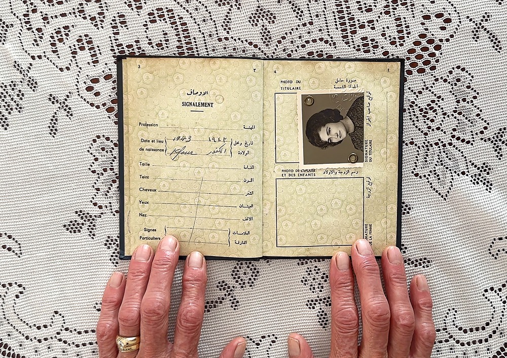 A photo showing an elderly woman's hand holding open an old pocket-sized book against a lace tablecloth. Arabic and French writing indicates it’s an identification document, with a younger woman’s photo on one page.