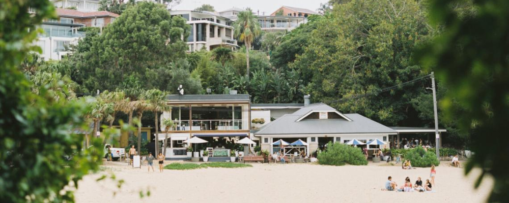 a two store venue that opens up onto the beach. there are people sitting at the venue and on the beach.